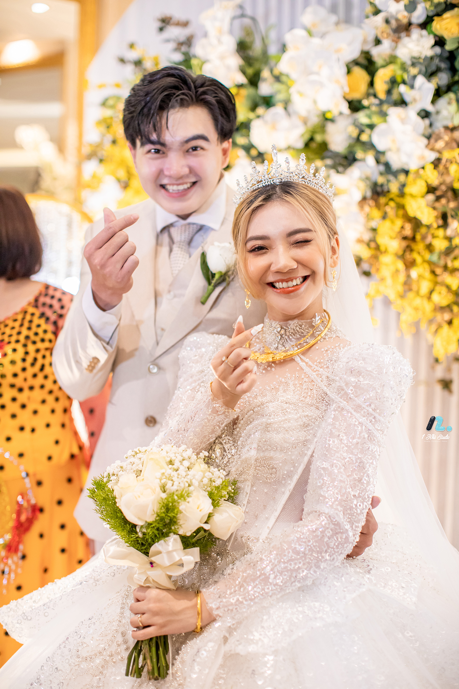 Cô dâu chú rể rạng rỡ làm cử chỉ bắn tim hạnh phúc trong ngày tổ chức tiệc cưới tại sảnh nhà hàng