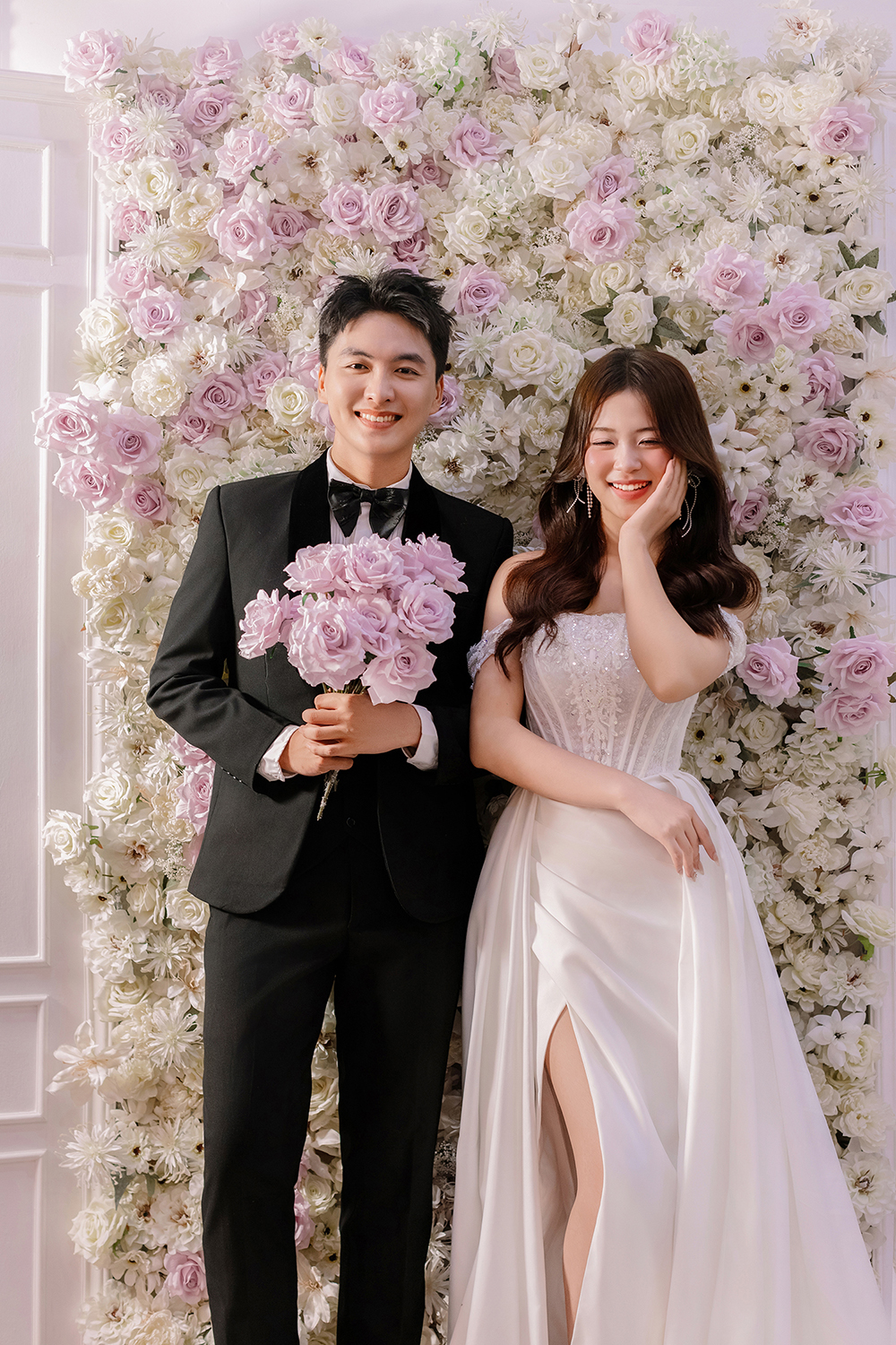 Phong cách chụp ảnh cưới Hàn Quốc trước phông hoa trong studio, cô dâu váy trắng và chú rể vest đen